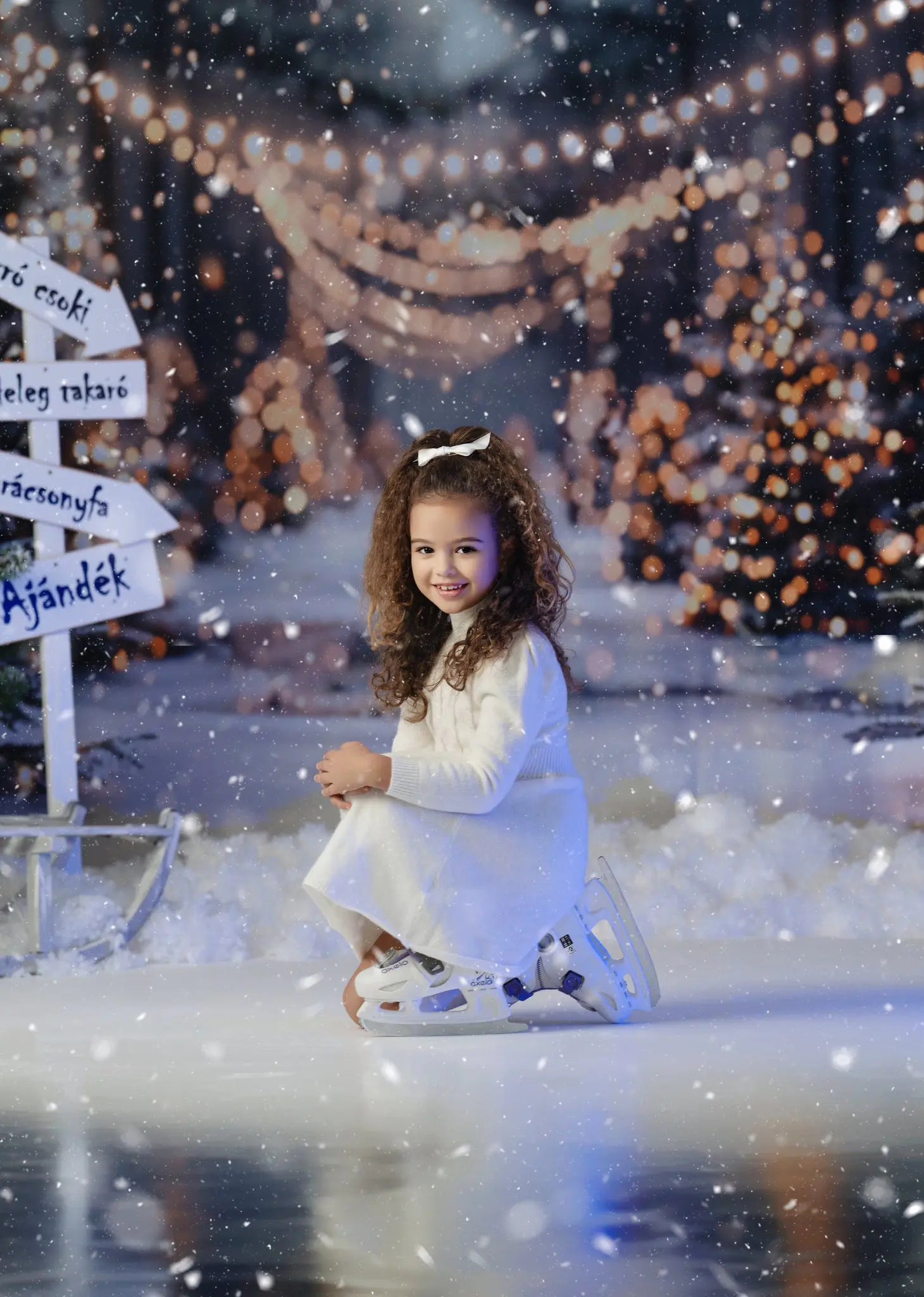 Young girl in a white dress sitting on a snowy ground with festive lights in a Christmas tree background