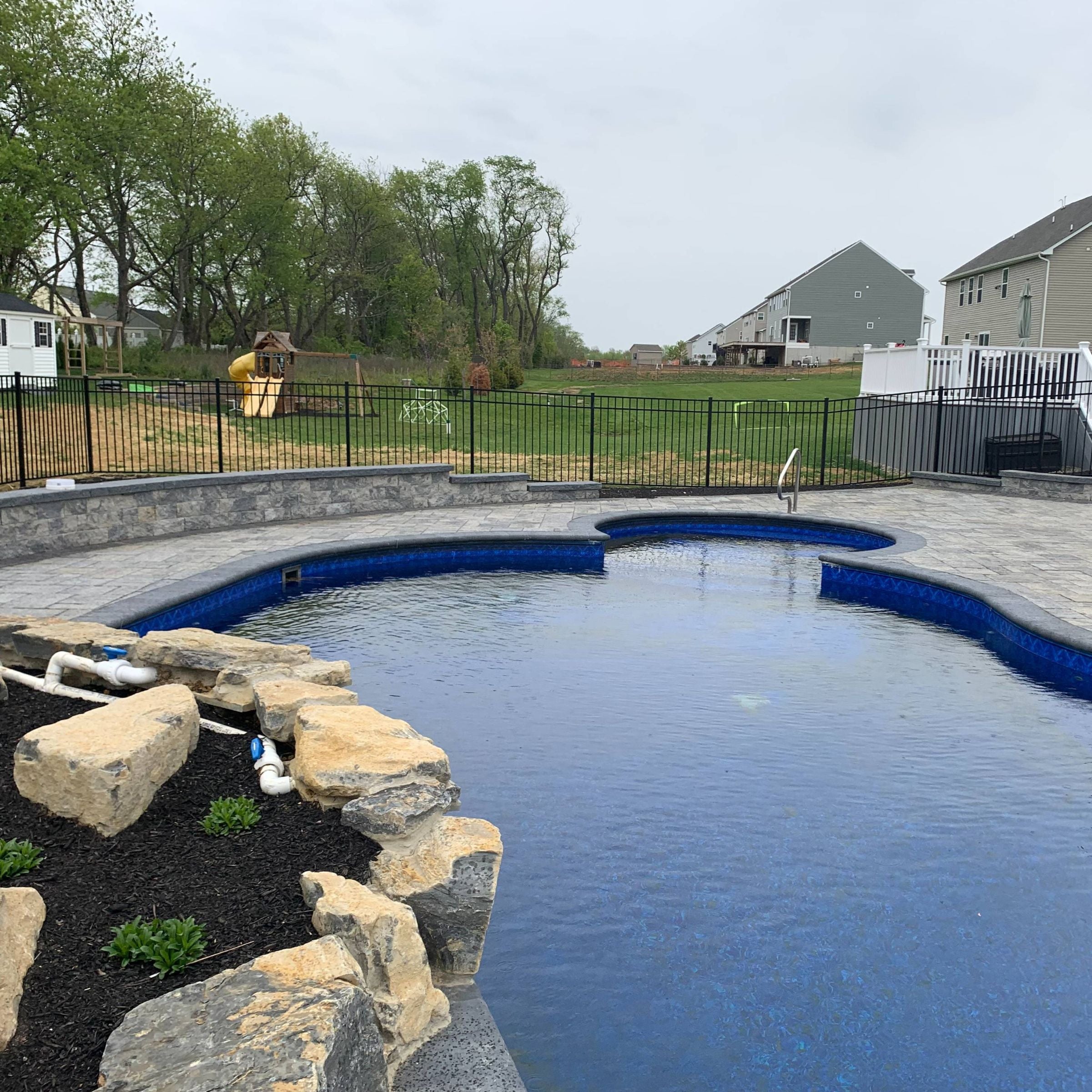 In-ground swimming pool featuring large natural boulder accents and a black-mulched garden bed, surrounded by a tiered stone retaining wall and grey paver decking.
