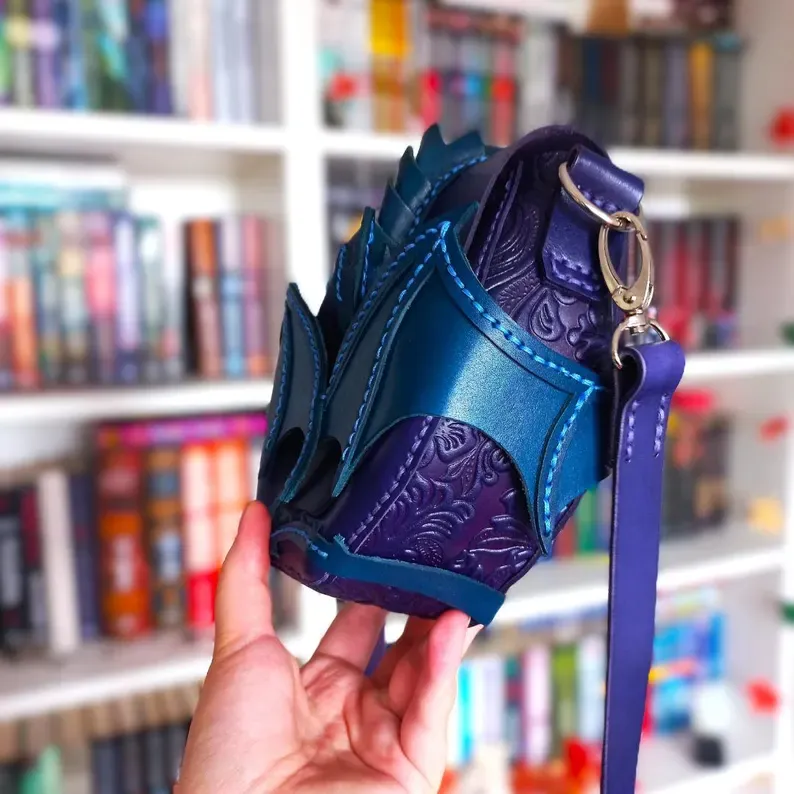 May include: A handcrafted, fantasy-themed crossbody bag in shades of blue and purple. The bag features a unique design with a dragon scale-like texture on the front, complemented by intricate embossed patterns. It has a long, adjustable strap and a silver-colored clasp.