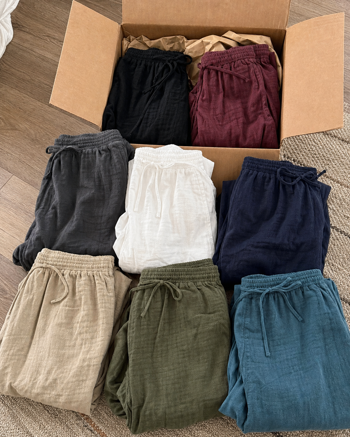 Eight separate wide-leg drawstring pants in black, burgundy, charcoal gray, white, navy, khaki, olive green and teal blue beside an unbranded box