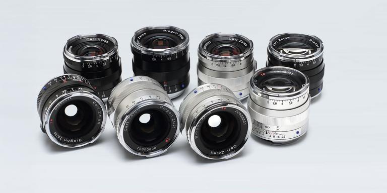 A collection of nine ZEISS camera lenses in black and silver finishes arranged on a light gray background.