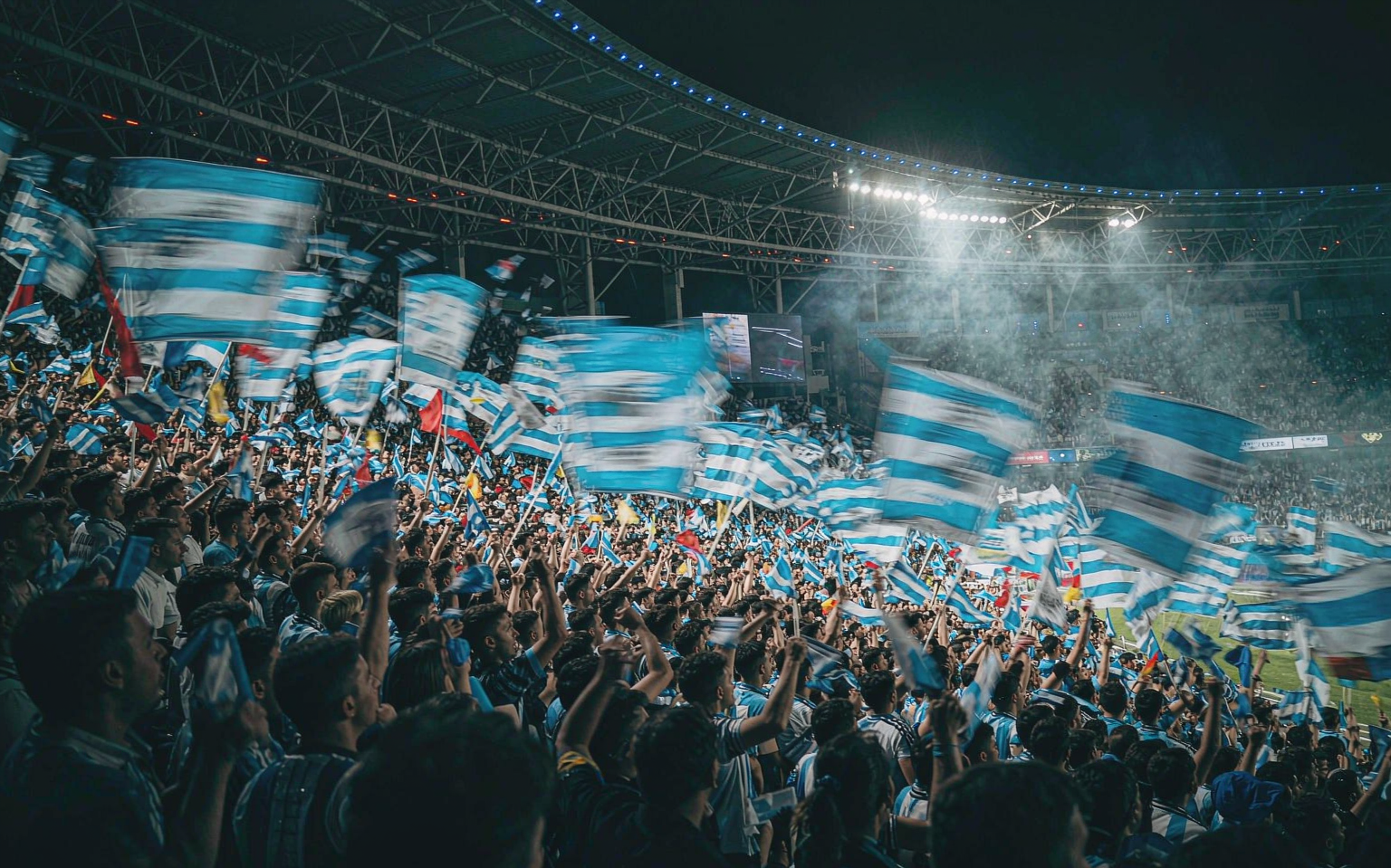 A floodlit stadium stand at night, fans waving sky blue and white flags