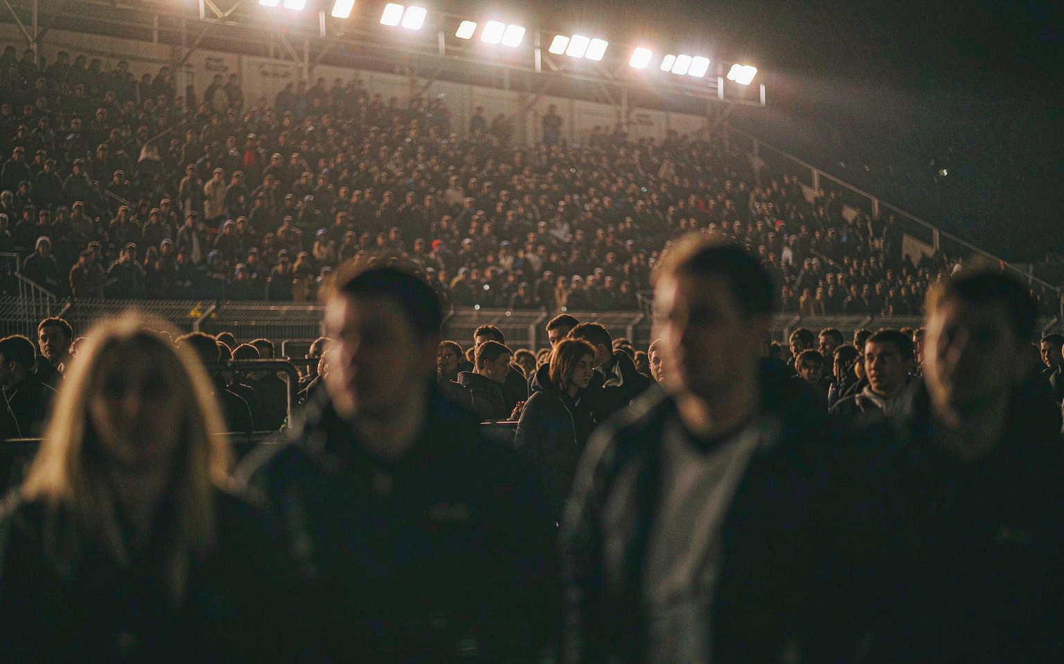 A floodlit stadium stand at night, blurred faces in the crowd, atmospheric haze and warm floodlight lens flares
