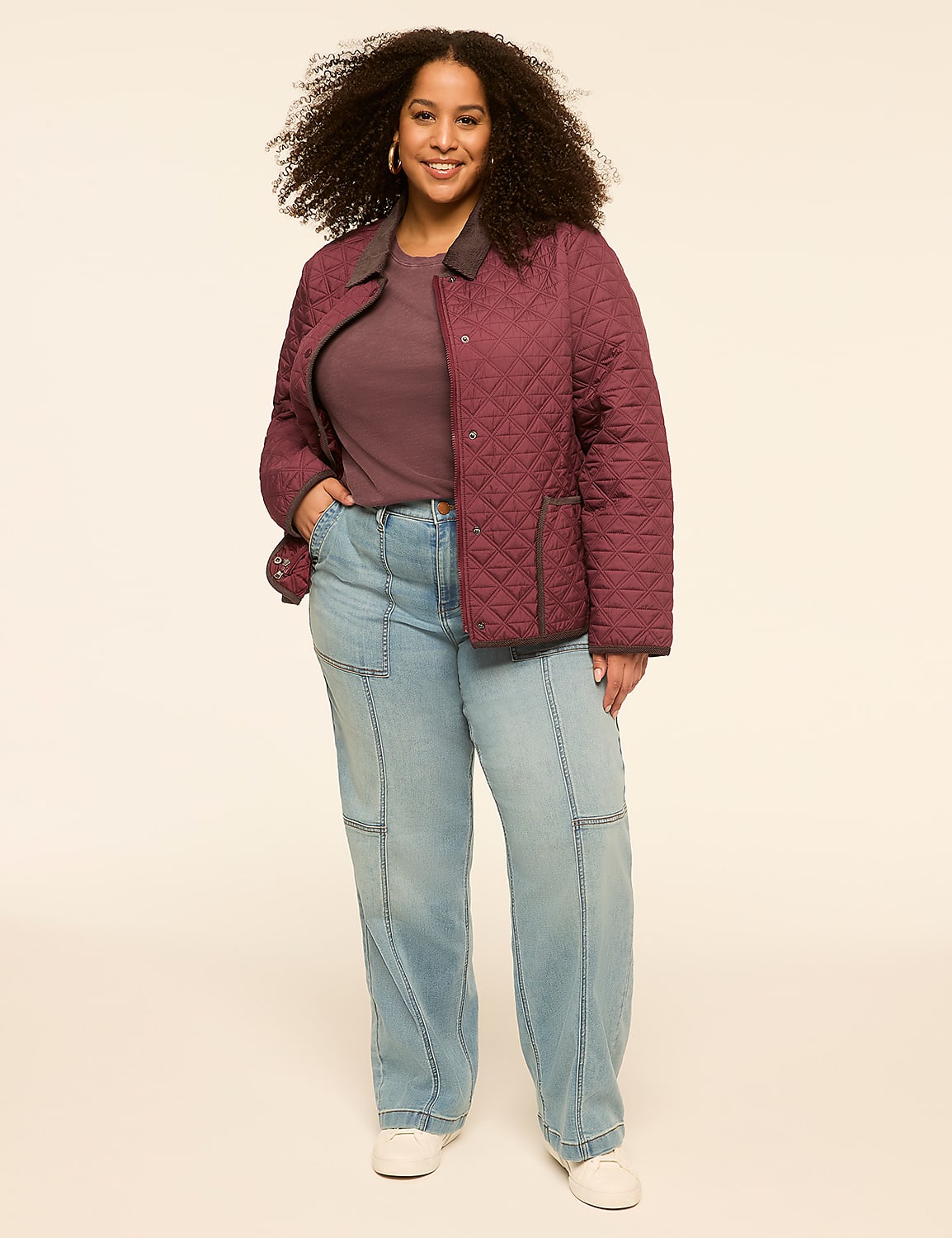 A smiling woman with curly hair stands in the foreground wearing a quilted burgundy jacket and light wash jeans against a plain, light-colored background. Product Image 3