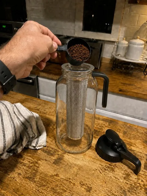 Coffee grounds being added to a stainless-steel infuser supported by its rim on the pitcher opening