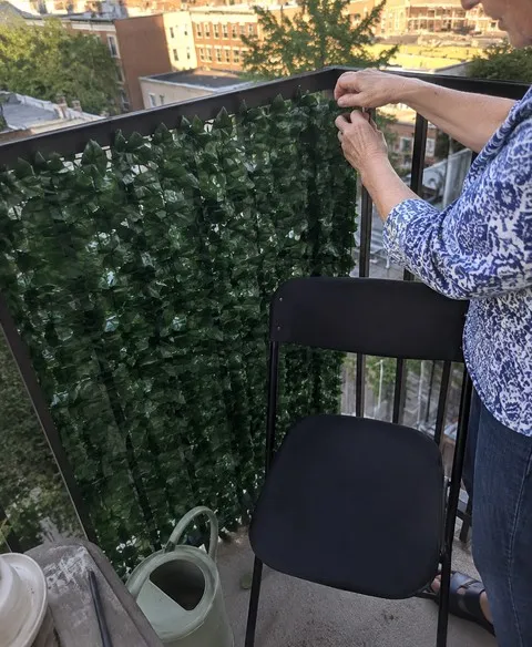 Older homeowner adjusting an artificial ivy privacy screen on a small balcony railing