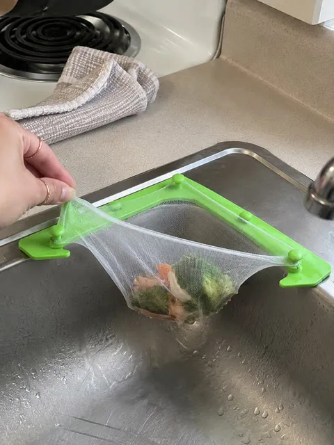 Hand lifting a mesh bag with vegetable scraps from a green sink holder