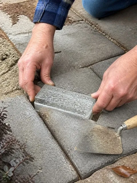 Hands setting a glass brick light into a backyard paver opening