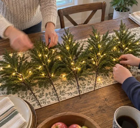 Family hands shaping five glowing pine stems across a dining table