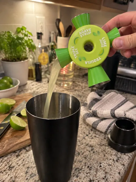 Four-way cocktail jigger pouring lime juice into a shaker on a home kitchen counter