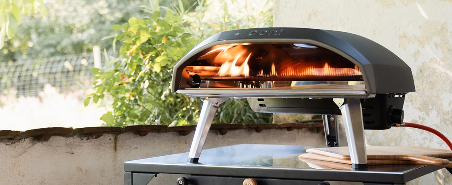 Portable outdoor pizza oven with dome-shaped black top showing active flames inside, positioned on a dark metal stand with wooden handle.