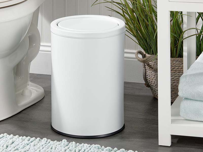 round swing top lid operated waste can in white next to toilet and white shelf in bathroom