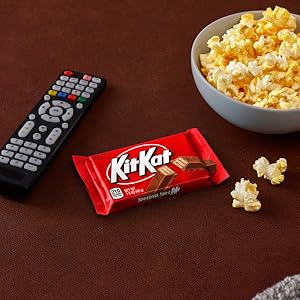 A Kit Kat candy bar placed between a television remote and bowl of popcorn