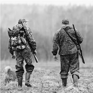 Black and white photo of 2 men in camo gear walking in a field with rifles slung on their backs.