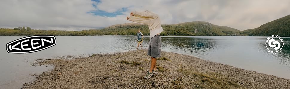 A man and a woman wearing KEEN sandals stand on the shore of a lake with grassy mountains across
