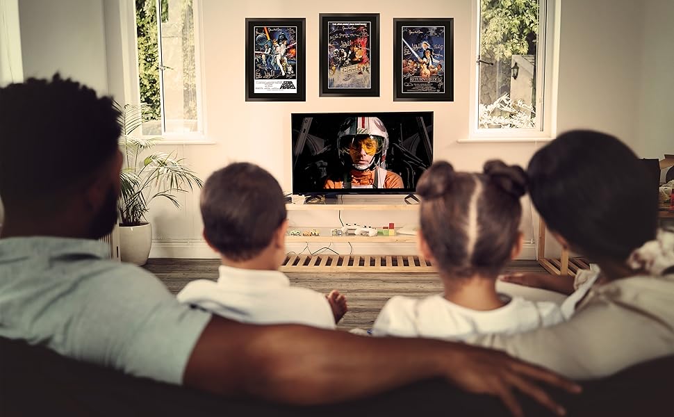 Living room scene showing people seated on a couch viewing a screen, with three framed artworks mounted on the wall above.