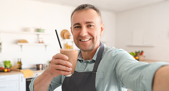 Smiling middle aged man taking a selfie while wearing an apron and holding a smoothie.