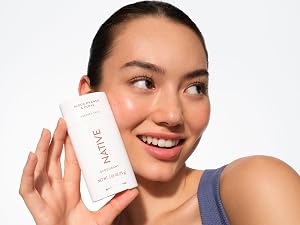 Smiling woman with fresh skin holds Native Blood Orange &amp;amp;amp; Clove deodorant, looking confident.