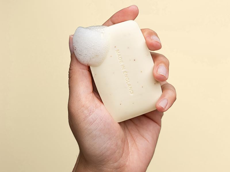 Cream-colored rectangular bar soap held in a human hand against beige background. Product shows smooth texture with subtle speckled pattern throughout.