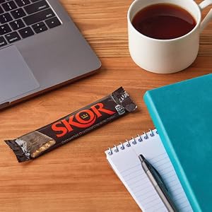 Full size Skor candy bar on a desk near a laptop, coffee mug, folders and a pen