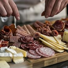 Hands arranging charcuterie board with rolled meat slices, cheese varieties, and garnishes on wooden surface.