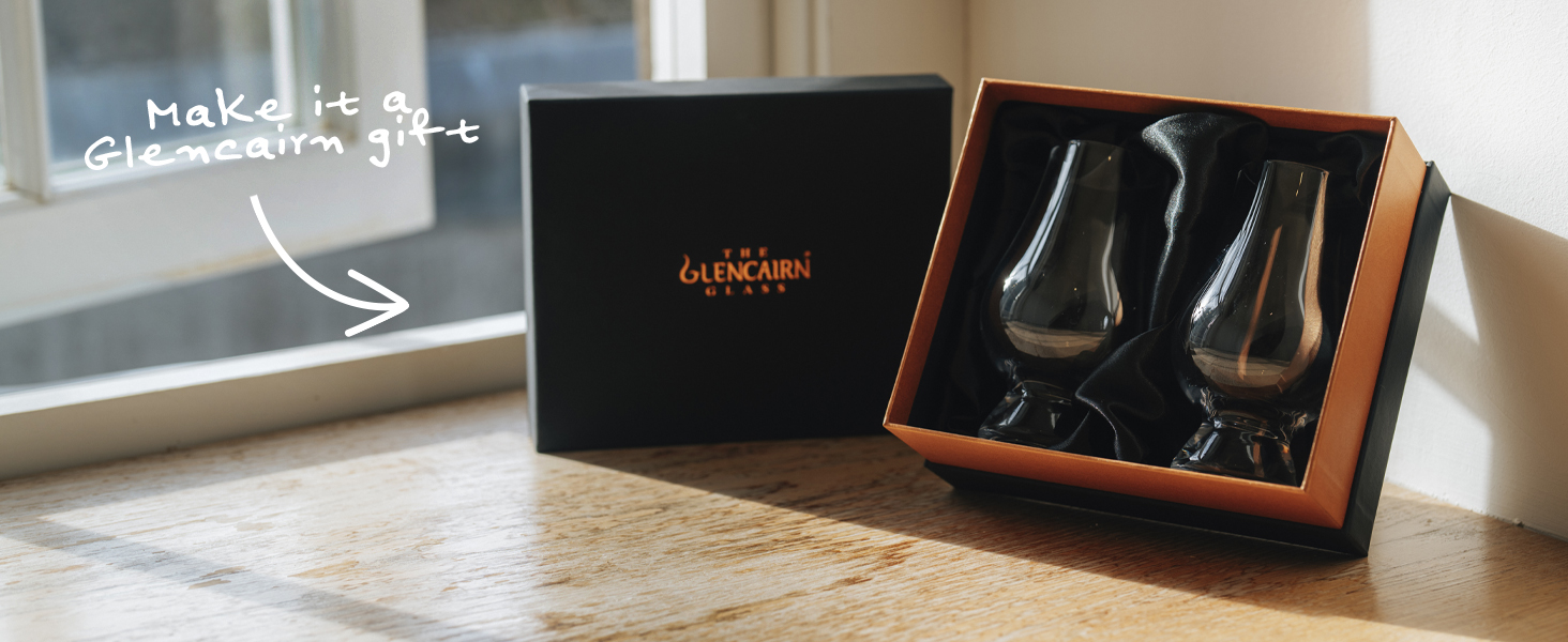 Open gift box containing two tulip-shaped whiskey glasses on a wooden surface. Black box with gold lettering visible nearby.