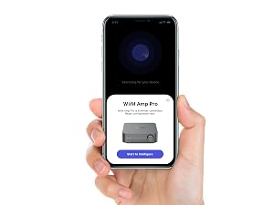 Hand holding smartphone displaying setup screen for WIIM Amp Pro device. Screen shows image of gray rectangular device and blue setup button.