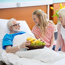 An elderly man in a hospital bed smiles at a young girl holding a basket of fruit. The man wears a blue gown, and the girl has on a red and white striped shirt.