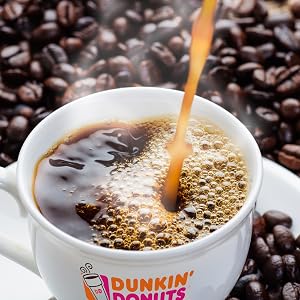Coffee being poured into a Dunkin Donuts mug resting on roasted coffee beans