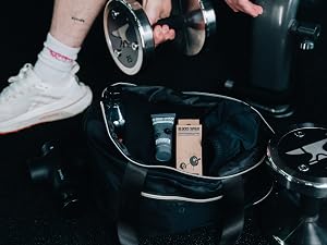 Fitness equipment including chrome dumbbells and a black gym bag containing supplements. A person's hands and legs are visible, handling the weights.