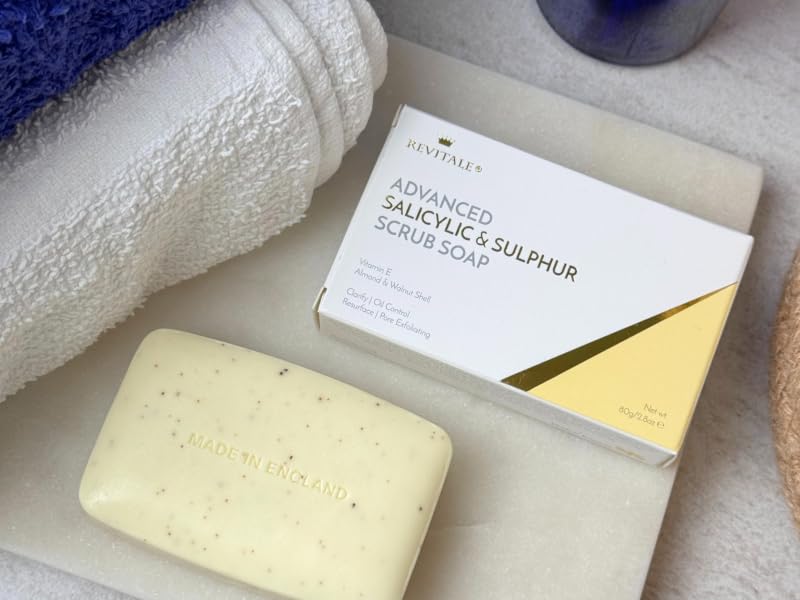 Text reads 'SULFURED SULFUR SOAP'. Cream-colored rectangular soap bar with product packaging displayed on white towel next to blue rolled cloth and skincare items.