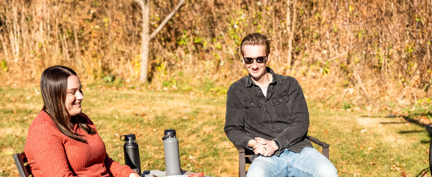 man and woman sitting in front of fire pit watching hotdogs and burgers cook on cooking grate
