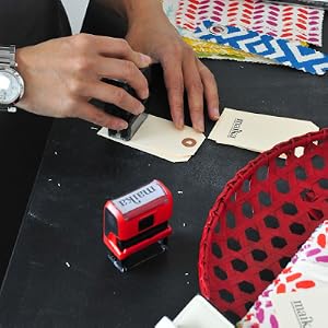 A hand stamps the MAIKA logo onto paper next to a pile of MAIKA fabric and pattern samples.