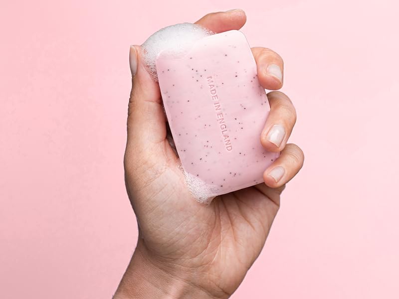 Person's hand holding small pink rectangular object against pink background, appears to be compact item held in palm.