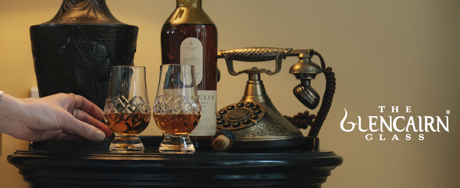 Two crystal whisky glasses with amber liquid, alongside a whisky bottle and vintage telephone on a dark wooden tray. Glencairn Glass logo visible.