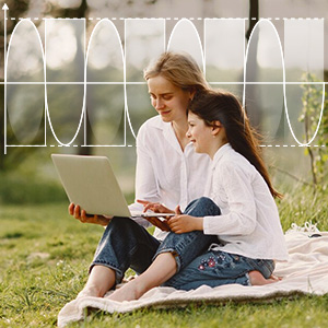 Two women sitting on grass, using laptop and mobile device. They wear white tops and are in an outdoor setting with a fence visible.