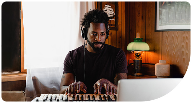Person sitting at a desk with headphones, playing an electric keyboard or MIDI controller in a dimly lit room with a lamp.