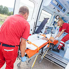 Emergency medical personnel in red uniforms attending to a patient on a stretcher near an emergency vehicle outdoors.