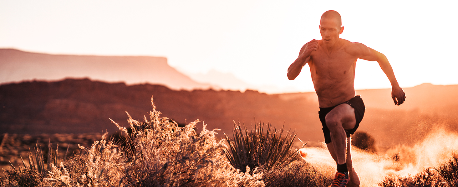 Shirtless man running through the desert at sunset, kicking up clouds of dust behind him.