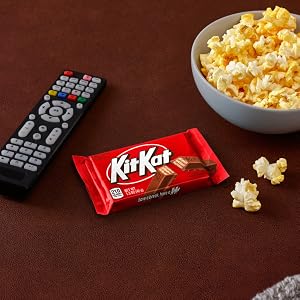 A Kit Kat candy bar placed between a television remote and bowl of popcorn