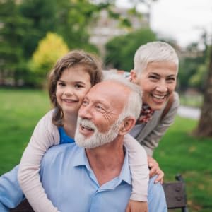 An older couple with white hair smiles while a young girl hugs both from behind. They are in a park