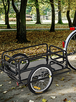 A bicycle trailer with metal frame and two wheels, parked on leaf-covered ground in a park with trees.