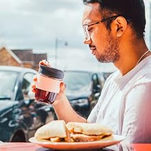 Man in pink shirt holding red beverage cup sitting outdoors with pizza and blurred urban background with cars and buildings.