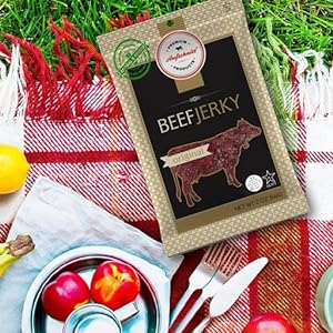 beef jerky on a picnic blanket next to a plate and cup with fruits. grass in background
