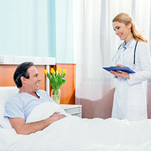 Medical consultation scene in a bright hospital room with medical professional holding clipboard beside patient in bed.