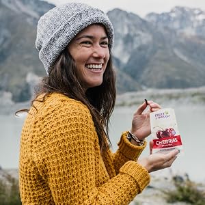 A woman smiles as she eats dried cherries next to a lake in the mountains.