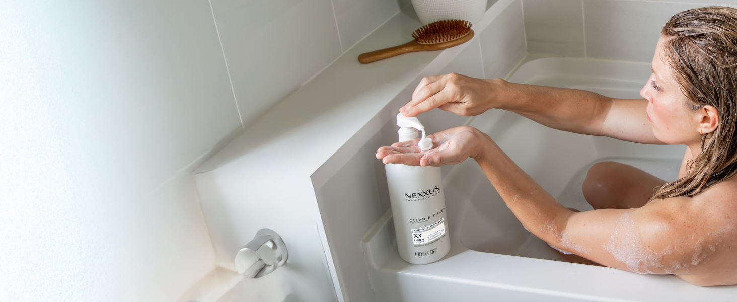 Woman in bath tub pressing bottle's pump and pouring Nexxus conditioner in hand.