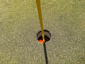 Golf hole on green with yellow flag pole. Orange golf ball visible at bottom of hole. Surrounding grass appears to be artificial turf.