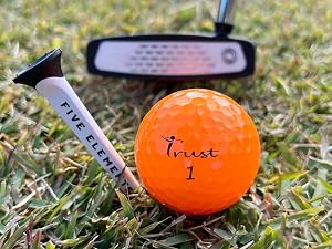 Orange golf ball labeled 'Trust 1' on grass, with a golf tee and putter visible in the frame.