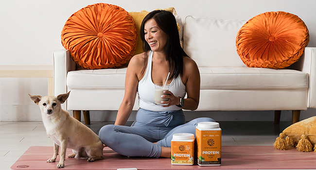 Young woman holding a smoothie sitting an a yoga mat with her dog next to Sunwarrior Classic Plus